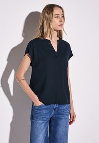 Top nero a maniche corte con scollatura a V, realizzato in tessuto morbido, caratterizzato da una vestibilità rilassata e semplici dettagli di cucitura. Abbinato a jeans blu.