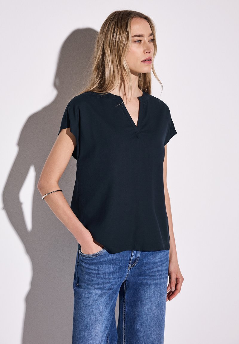 Top nero a maniche corte con scollatura a V, realizzato in tessuto morbido, caratterizzato da una vestibilità rilassata e semplici dettagli di cucitura. Abbinato a jeans blu.