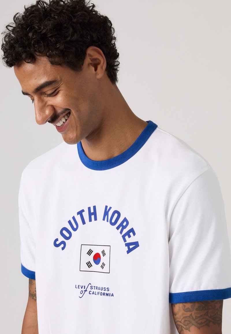 Homme aux cheveux bouclés portant un t-shirt blanc avec un liseré bleu, texte South Korea, drapeau et logo Levi Strauss of California, souriant et regardant vers le bas.