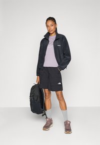 Fleece jacket in navy, purple T-shirt, black cargo shorts with logo, gray socks, and brown hiking shoes; carrying a black backpack.