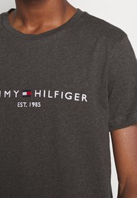 Tmavě šedé bavlněné tričko s bílým vyšívaným logem "TOMMY HILFIGER" a malým červeným a modrým obdélníkem, kulatým výstřihem a krátkými rukávy.