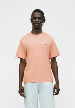 Hombre de piel oscura y cabello trenzado que lleva una camiseta de manga corta color durazno con un pequeño logo y jeans azul claro, de pie contra un fondo blanco.