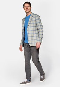 Jasnoszary marynarka w kratkę z niebieskimi akcentami na granatowej koszulce, zestawiona z dopasowanymi szarymi dżinsami i szarymi trampkami, idąca na jednolitym tle.