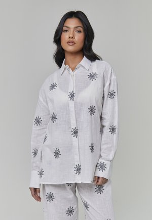 Hvid linskjorte med sort solbroderi, knaplukning foran og lange ærmer. Har en afslappet pasform og diskrete teksturer over det hele.