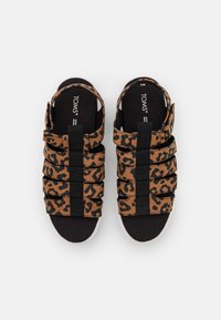 Sandálias com padrão de leopardo castanho, com tiras elásticas pretas e solas brancas. Palmilha acolchoada macia para conforto. Nome da marca "TOMS" visível no interior.