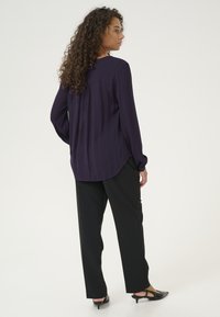 Blusa lunga a maniche con un fit rilassato, di colore viola scuro, in tessuto strutturato e con orlo curvo, abbinata a pantaloni neri a gamba dritta e tacchi neri.