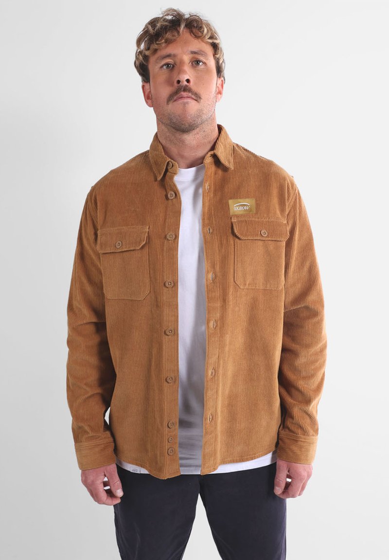 Veste en velours côtelé marron avec deux poches poitrine, fermeture par boutons et un petit patch de marque sur le côté gauche. Présente une coupe décontractée et un tissu texturé.