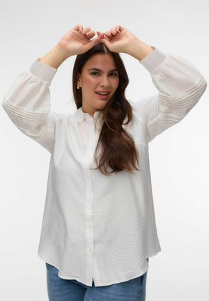 Femme aux longs cheveux bruns portant un chemisier blanc et un jean bleu, levant les deux mains au-dessus de sa tête avec les doigts touchés.