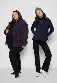 Duas mulheres estão lado a lado. À esquerda: casaco multicolorido e peludo, blusa preta, calças pretas, sapatos azuis. À direita: blusa azul-marinho com fecho, calças pretas, sapatos brancos.