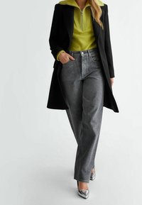 Cappotto nero su un maglione verde brillante con colletto a zip, abbinato a pantaloni in denim grigio con dettagli argentati e tacchi alti argentati lucidi.