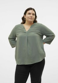 Blouse légère vert olive avec un décolleté en V, des manches 3/4, une texture subtile et une coupe décontractée, portée avec un pantalon noir.