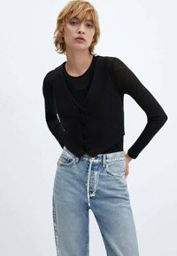 Zwarte vest met een V-hals en vijf knopen, gedragen over een zwart shirt met lange mouwen, gecombineerd met lichtblauwe hoge taille jeans.