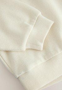Krämfärgad sweatshirt med ribbade muddar och nederkant, med en mjuk, texturerad yta och en enkel design utan mönster eller detaljer.