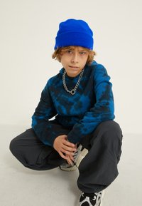 Felpa blu tie-dye con maniche lunghe, abbinata a pantaloni neri larghi. Un berretto di lana blu e una collana in argento completano il look.