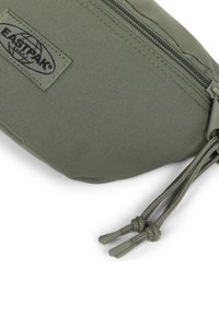 Bolsa de nylon verde con cierre de cremallera, que presenta una superficie texturizada, parche con el logo de Eastpak y dos tiradores de cremallera de cordón.