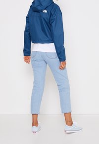 Niebieska krótka kurtka przeciwdeszczowa z kapturem, jasnoniebieskie dżinsy i jasnoniebieskie trampki. Widoczny logotyp marki na kurtce. Prosty i swobodny wygląd.