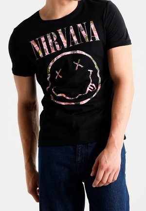 Persoon draagt een zwart T-shirt met de tekst "NIRVANA" en een roze smiley met bloemenpatroon, gecombineerd met donkerblauwe spijkerbroek.