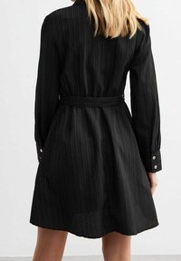 Abito camicia nero realizzato in tessuto voluminoso, con vita cintata, polsini abbottonati e orlo a ginocchio.
