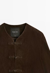 Vêtement en daim marron foncé avec un col rond et trois fermetures horizontales à boucles et nœuds sur le devant, étiquette à l'intérieur du col.