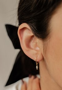 Orecchino a cerchio d'oro con una perla bianca pendente e un piccolo accento di pallina d'oro, esposto vicino all'orecchio di una donna e abbinato a lunghi capelli scuri.