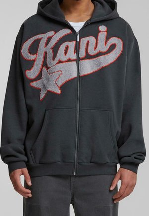 Person trägt einen dunklen Kapuzenpullover mit Reißverschluss mit großem strukturiertem „Kani“-Schriftzug und Stern-Motiv in Grau, das rot umrandet ist, auf der Brust.