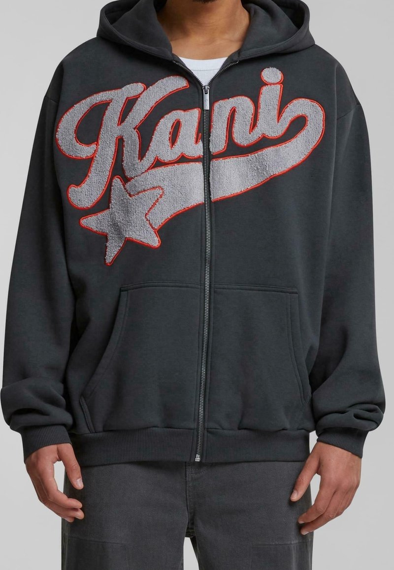 Personne portant un sweat à capuche zippé sombre avec un grand texte « Kani » texturé et un motif étoile gris avec un contour rouge sur la poitrine.