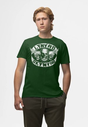 Jeune homme aux cheveux blonds portant un t-shirt vert foncé Lynyrd Skynyrd et un pantalon foncé, debout avec une main dans la poche.
