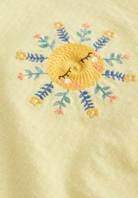 Un tissu jaune présente une broderie de soleil avec des yeux fermés, entourée de motifs floraux en bleu, rose et blanc, avec une finition texturée.