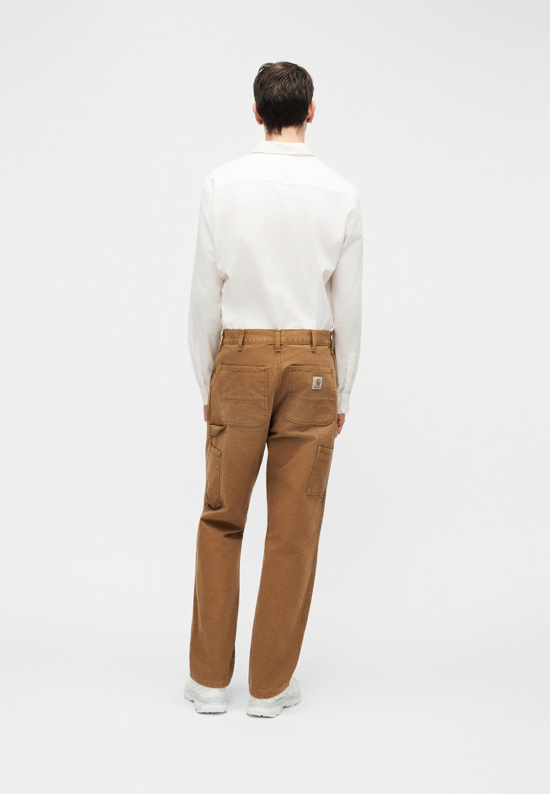 Homme portant une chemise blanche à manches longues, un pantalon cargo marron avec poches arrière et patch logo, ainsi que des baskets blanches, debout de dos.