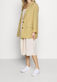 Żółta marynarka w kratkę o luźnym kroju, zapinana na jeden guzik, w połączeniu z jasnobeżową sukienką i białymi sneakersami. Gładka tekstura materiału.