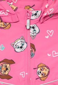 Veste rose zippée avec capuche, ornée de visages de chiens de dessin animé et de cœurs de différentes couleurs. Le matériau semble lisse et léger.