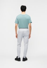 Άνδρας που στέκεται με την πλάτη στραμμένη, φοράει ανοιχτό μπλε κοντομάνικο πουκάμισο, ανοιχτό γκρι φόρμα, άσπρες κάλτσες και μαύρα αθλητικά σε λευκό φόντο.