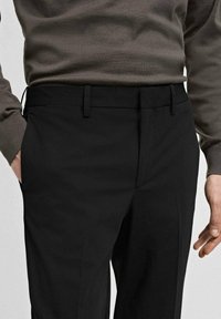 Schwarze Hosen aus glattem Stoff, mit geradem Schnitt, Gürtelschlaufen und Seitentaschen. Getragen mit einem taillierten, strukturierten braunen Pullover.