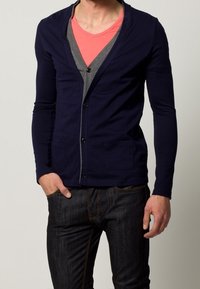 Cardigan lungo blu navy con maniche lunghe, bottoni frontali, bordi grigi e due tasche frontali, indossato sopra una maglietta rosa a V.