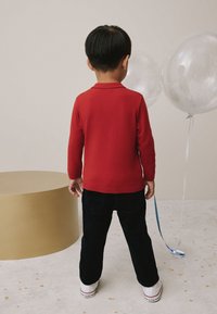 Rode trui met lange mouwen, marineblauwe broek, witte schoenen. Kind met de rug naar de kijker, houdt een lint van een blauwe ballon vast, met een gouden ronde tafel op de achtergrond.