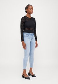 Crna majica dugih rukava od prozirnog teksturiranog materijala, u kombinaciji s svijetloslikaškim skinny trapericama i crnim slingback cipelama s ukrasnim hardverom.