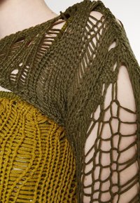 Olivengrøn strikket beklædning med indviklede mønstre; har åbent vævning og teksturerede designs. Inkluderer et gult lag nederst, der er synligt under.