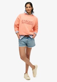 Sudadera de color coral con "J'ADORE SUPERDRY" en letras grandes, superpuesta sobre una camisa de color azul claro, combinada con pantalones cortos de denim y zapatillas beige.