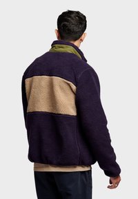 Purple fleece jacket with a tan horizontal band across the back, featuring a green collar, textured surface, and relaxed fit.