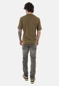 Homme aux cheveux bruns et bouclés, de dos, portant un t-shirt vert olive, un jean gris, des baskets Nike, avec un tatouage visible sur le bras gauche.