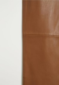 Gros plan d'un vêtement en cuir marron montrant une texture lisse avec des coutures visibles sur un fond clair uni.