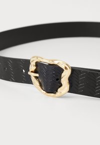 Črn pas iz teksturiranega materiala z zlatim, nepravilno oblikovanim sponko in herringbone vzorcem na pasu.