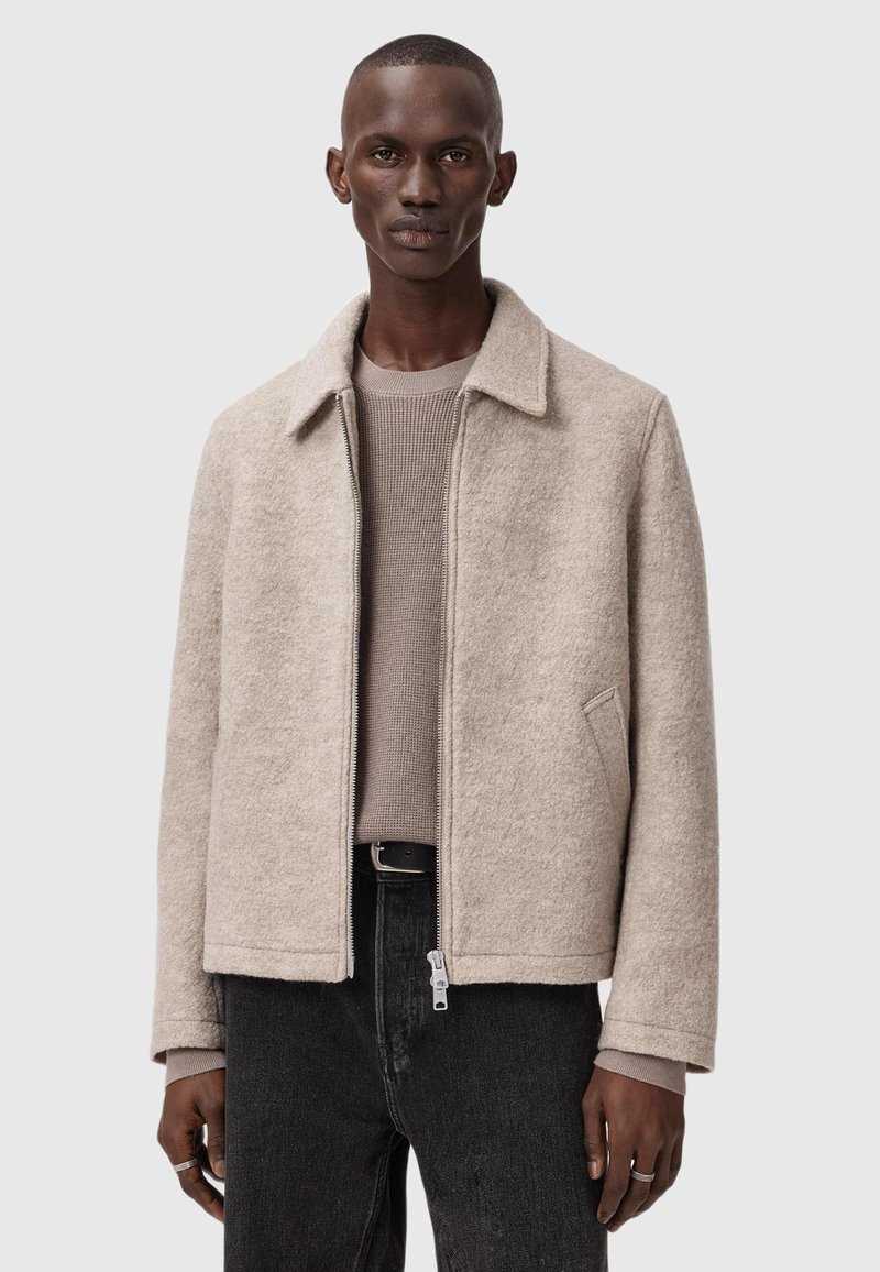 Beige uldblandingsjakke med kravesnit, lynlås lukning og to sidelommer, parret med en lys brun struktureret sweater og sorte jeans.