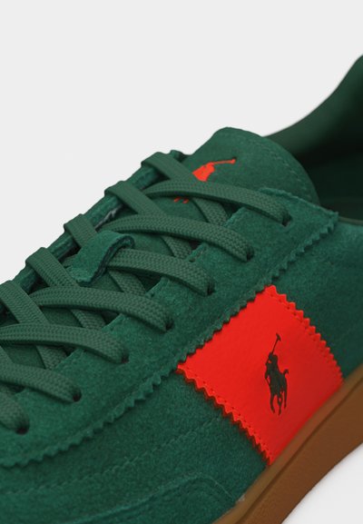 Πράσινο σουέντ αθλητικό παπούτσι με κορδόνια στο ίδιο χρώμα, μια φωτεινή πορτοκαλί πινακίδα με μαύρο λογότυπο παίκτη πόλο και καφέ λαστιχένια σόλα.