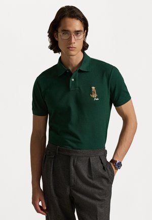 Zielona koszulka polo z krótkim rękawem, z haftowanym logo psa i napisem "Polo." Dobrze dopasowane szare spodnie.