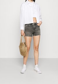 Nainen, jolla on päällä valkoinen lyhyt nappipaita, valkoinen toppi, harmaat denimit, valkoiset korkeavartiset sneakersit ja joka pitää käsissään pyöreää punottua käsilaukkua.