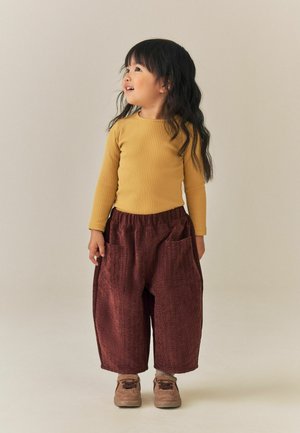 Junges Kind mit langen dunklen Haaren, das ein senfgelbes Oberteil, lässige bordeauxrote Hose und braune Schuhe trägt und zur Seite schaut.