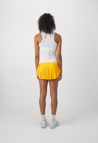 White sleeveless top with a keyhole back, paired with a pleated yellow skirt. Model wearing gray sneakers and crew-length socks.