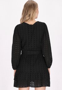 Schwarzes Langarmkleid mit Durchbrüchen, tailliertem Schnitt und gestuftem Rock. Der Stoff hat eine weiche Textur mit Blumenmustern.