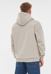 Hanorac bej cu glugă, cu o croială lejeră, manșete și tiv, cutate; textură netedă a materialului. Purtați peste blugi albaștri.